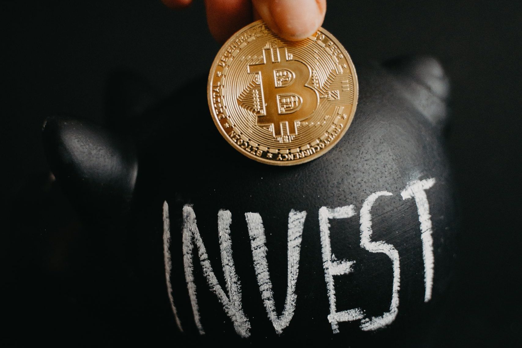 Gold bitcoin inserted into a black piggy bank labeled 'Invest', symbolizing cryptocurrency saving.
